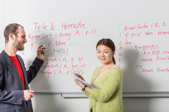 Two people in front of a whiteboard. One is holding a red pen, the other a notepad.