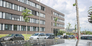 Informatik Informatikgebäude: Ein großes, braun-rot verklinkertes Gebäude mit großen Fenstern und Flachdach. Davor auf der Straße parken Autos hintereinander.